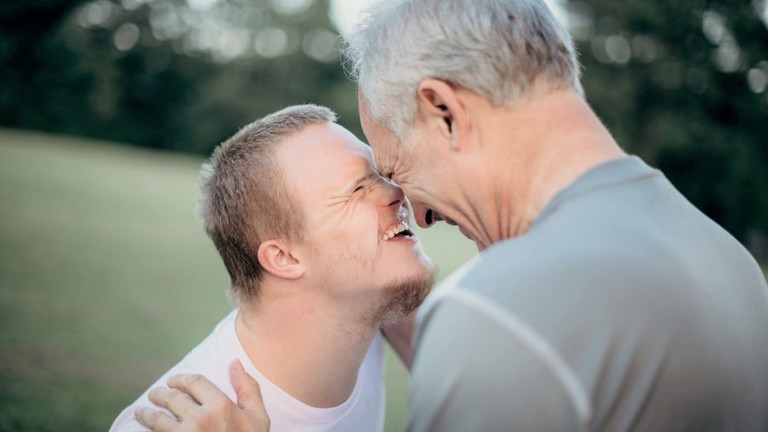 Vater mit grauem Haar berührt mit der Stirn seinen etwa 20jährigen Sohn mit Trisomie 21, beide lachen herzlich