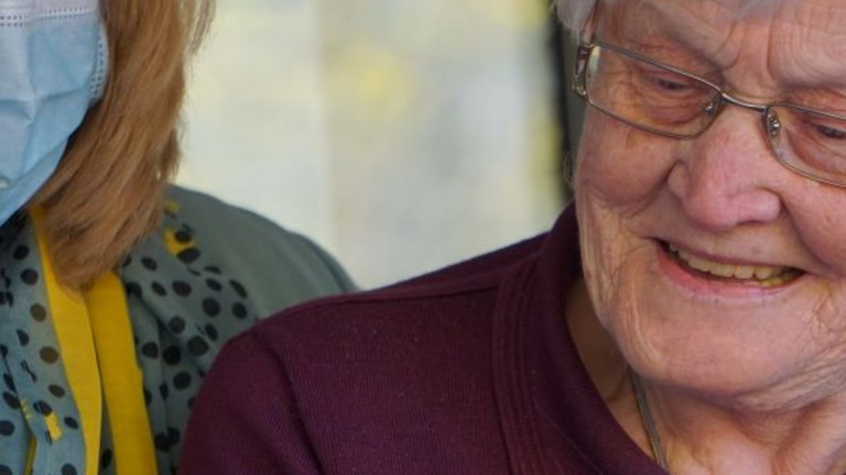Ältere Frau blickt fröhlich gemeinsam mit einer jüngeren Frau und Mundschutz auf ein Tablett-Computer