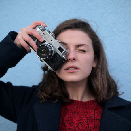 junge Frau vor blauer Wand hält sich eine alte Kamera vor ein Auge 