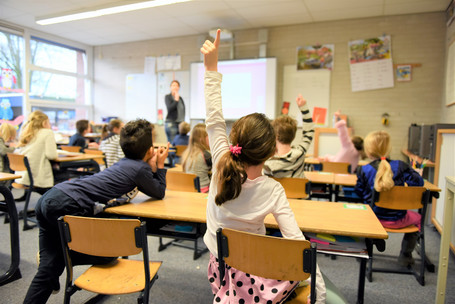 Ein Klassenraum mit Grundschul-Kindern von hinten zur Tafel hin fotografiert. Ein dunkelhaariges Mädchen in der letzten Bank meldet sich.