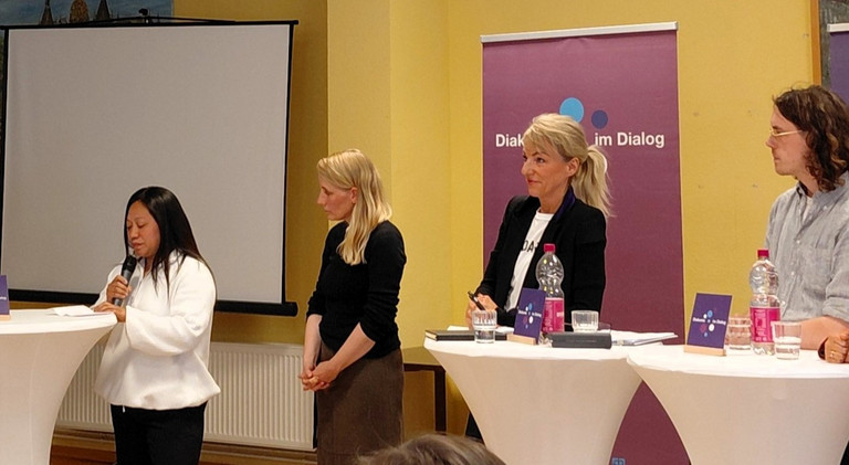 Terryflor, Pflegekraft von den Philippinen spricht auf dem Podium über ihr Ankommen in Schwerin. Politiker:innen und Moderatorin hören interressiert zu