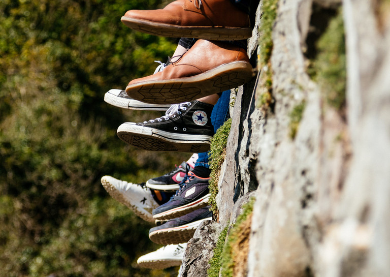 4 Paar Füsse in unterschiedlichen Schuhen  von Menschen, die auf einer Gartenmauer sitzen
