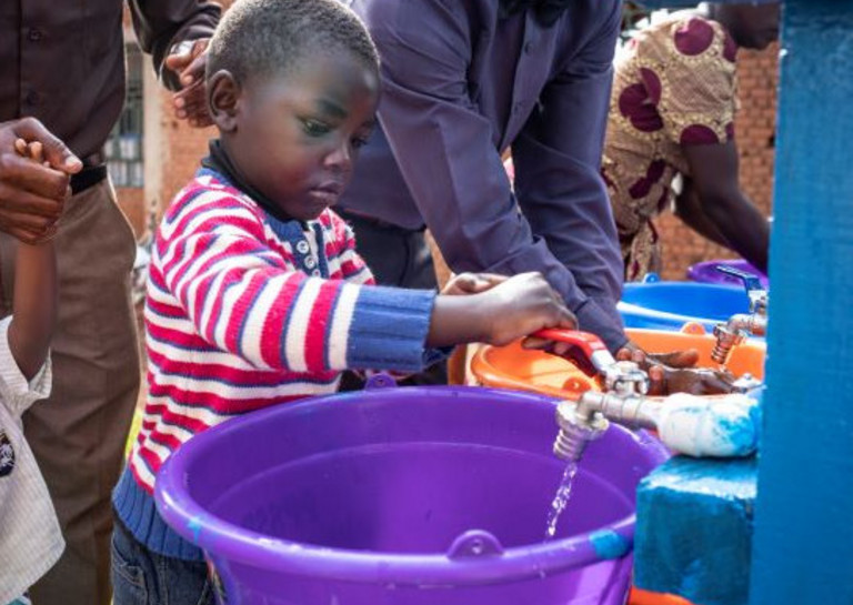 Junge im Vorschulalter in der DR Kongo mit rotgeringeltem Pulli wäscht sich die Hände über violetter Schüssel